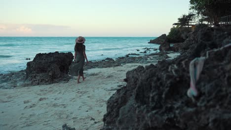 Woman-is-slowly-walking-on-the-beach-near-rocks-on-gently-sea-breeze,-wearing-a-dress-and-sunhat