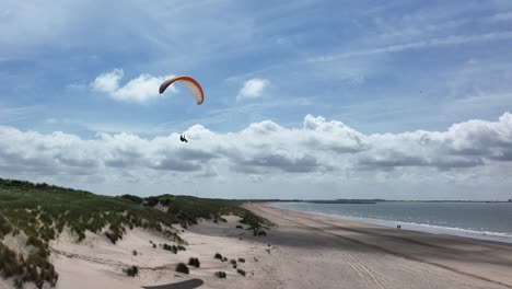 Drone-backing-away-from-paraglider-drifting-above-lush-seafront-sand-dunes