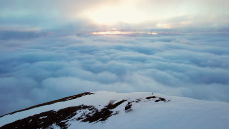 Toma-Aérea-Estática-Extremadamente-Larga-De-Un-Montañero-Parado-En-El-Pico-Cubierto-De-Nieve-De-Una-Montaña-Mirando-Sobre-Una-Increíble-Capa-De-Nubes-Con-El-Sol-Poniente-En-El-Horizonte,-Sol-De-Medianoche-Noruega