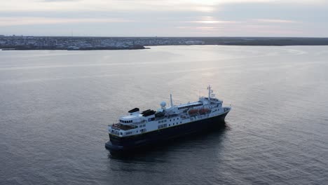 Expedition-cruise-ship-anchored-near-shore-of-Iceland-during-cloudy-sunset,-aerial