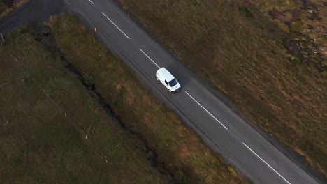 White-car-travels-on-small-rural-road-with-grass-on-sides,-cloudy-day,-tracking-aerial-shot