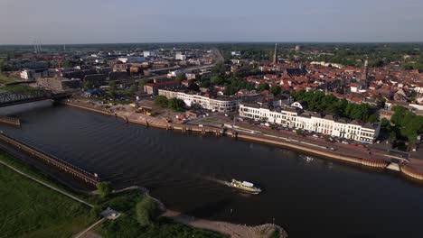 Aerial-following-of-inland-shipping-large-cargo-vessel-leaving-ripple-waves-on-river-IJssel-passing-IJsselkade-boulevard-countenance-cityscape-of-tower-town-Zutphen