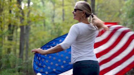 Pretty-blonde-woman-with-sunglasses-on-in-a-forest-spinning-around-to-the-right-with-a-US-flag-flying