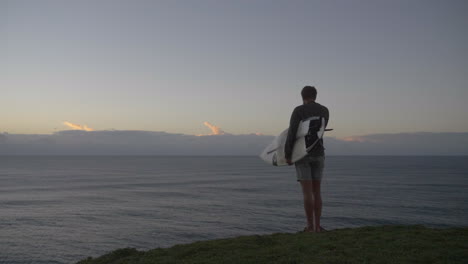Man-standing-on-edge-of-cliff-looking-at-ocean-at-sunrise-holding-surfboard