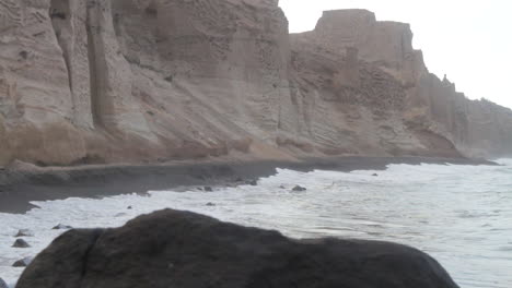 Tracking-shot-of-waves-hitting-a-black-beach-that's-surrounded-by-tall-white-volcanic-cliff-formations