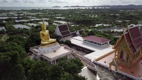 Un-Disparo-Orbital-Lento-Alrededor-De-Una-Estatua-De-Oro-De-Buda-Sentado-Y-Un-Templo,-Tailandia