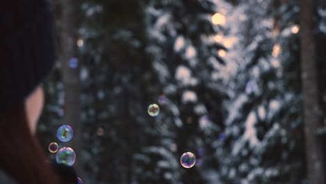 Brünette-Frau-Mit-Winterkleidung,-Die-Regenbogenfarbene-Seifenblasen-Bläst,-Die-In-Der-Luft-Schweben,-Mit-Einem-Verschneiten-Wald-Im-Hintergrund