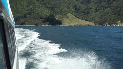 Foamy-waves-breaking-on-side-of-cruise-boat-in-Marlborough-Sounds,-New-Zealand-with-green-hills-in-background