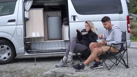 Young-attractive-couple-sitting-and-eating-lunch-with-motorhome-by-Lake-Wakatipu,-Queenstown,-New-Zealand
