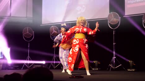 Zwei-Cos-spielerinnen-In-Anime-charakterkostümen,-Die-Japanische-Kimonos-Als-Fiktive-Charaktere-Auf-Der-Bühne-Beim-Spiel-Auf-Der-Expo-Vor-Dem-Publikum-Mit-Violetten-Laserlichtern-Tragen