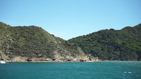 Shot-from-cruise-boat-of-beautiful-blue-sea-ocean-water,-green-hills-and-small-boat-in-Marlborough-Sounds,-New-Zealand