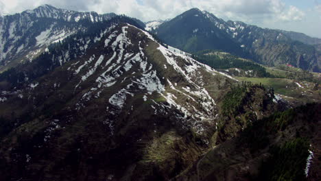 Antenne,-Die-über-Die-Eisigen-Berge-Zu-Den-Häusern-Der-Bergspitze-Führt,-Andere-Große-Bergwälder-Mit-Wolken-Und-Blauem-Himmel-Im-Hintergrund