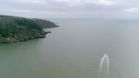 Aerial-landscape-shot-of-south-Devon-cliffs-with-a-wide-expanse-of-murky-ocean,-and-a-white-motorboat-sailing-out-to-open-sea