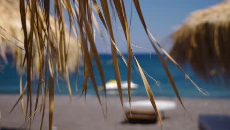 Handheld-Close-Up-of-Dry-Palm-Leaves-Moving-by-The-Wind-from-a-Tiki-Hut-Umbrella-on-The-Black-Lava-Beach-in-Perissa-Santorini-in-Greece-Europe