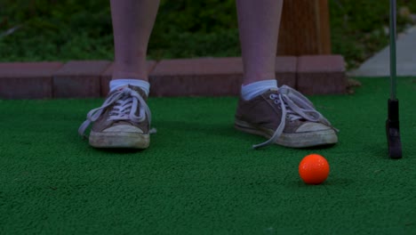 Ein-Golfer-Auf-Einem-Minigolfplatz,-Der-Einen-Orangefarbenen-Golfball-Auf-Den-Boden-Legt-Und-Sich-Bereit-Macht,-Den-Ball-Zu-Schießen