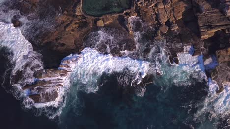 Top-bird's-eye-view-of-ocean-blue-waves-crashing-against-rocky-Australian-coastline-and-rockpool-drone-footage