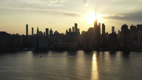 Luftdrohnenkamera-Schwenkt-Nach-Links,-Während-Ein-Boot-Mit-Einem-Goldenen-Sonnenuntergang-In-Die-Gleiche-Richtung-Schwimmt---New-York-City-Im-Hintergrund