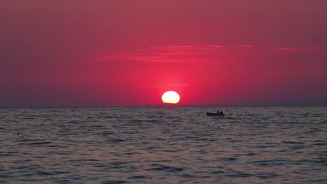 Red-golden-hour-and-the-sun-vanishing-beyond-the-horizon,-with-a-small-boat-passing-through