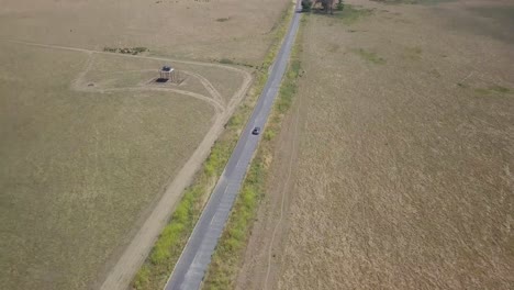 4K-Aerial-shot-of-silver-SUV-driving-down-long-straight-country-road,-isolated-against-wide-open-golden-desolate-fields-as-camera-pulls-back-and-reveals-wide-expanse-and-horizon-with-hills-and-sky