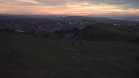 Lateral-tracking-shot-of-girl-running-on-the-Arthurs-seat-mountain-in-evening,-dusk-with-city-of-Edinburgh-lights-in-the-background-during-wonderul-sunset-light-and-blue-hour