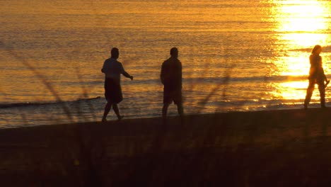 Eine-Frau-Und-Zwei-Männer,-Die-In-Der-Morgendämmerung-Am-Strand-Spazieren-Gehen,-Sonnenlicht-Auf-Dem-Wasser