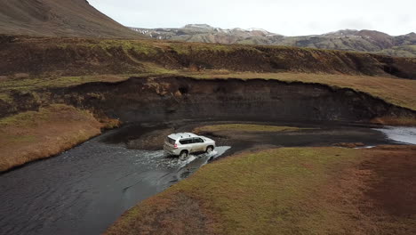 Four-Wheeler-on-Rough-Offroad-and-Shallow-River-in-Iceland-Countryside-Under-Volcanic-Hills,-Aerial-View