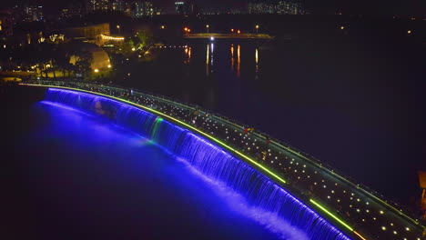 Wide-aerial-crane-shot-of-Starlight-Bridge-or-Anh-Sao-Bridge-at-night,-a-pedestrian-bridge-with-colored-lights-and-waterfall-in-District-7-of-Ho-Chi-Minh-City-or-Saigon,-Vietnam