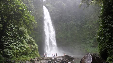 A-big-waterfall-crashing-down-on-big,-black-rocks-in-the-jungle-of-Bali-with-a-group-of-tourists-standing-and-watching-it