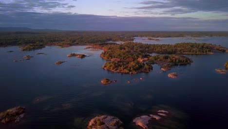 Rocky-Granite-Islands-with-Green-Pine-Trees-in-Clear-Blue-Lake-at-Sunset,-Drone-Aerial-Wide-Dolly-In