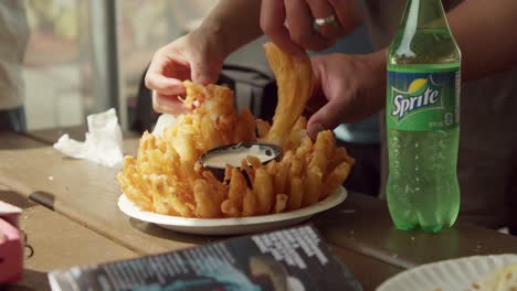 Hands-grabbing-onto-fried-blooming-onion-dish-with-dip-on-table-and-a-bottle-of-pop-at-the-side