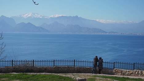 Full-shot,-couple-besides-the-railing,-cliffs-of-Antalya-turkey,-looking-at-the-scenic-view-of-the-Mediterranean-sea-and-toros-mountain-in-the-background