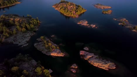 Rocky-Granite-Islands-with-Green-Pine-Trees-in-Blue-Lake-at-Sunset,-Drone-Aerial-Wide-Tilt-Reveal