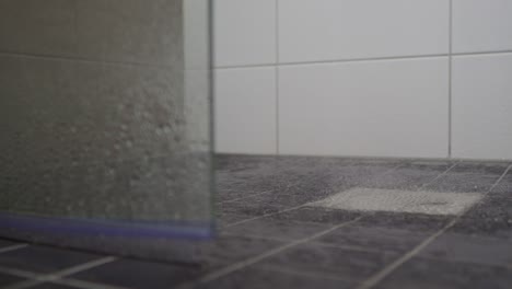 Water-droplets-splashing-on-bathroom-floor-into-drain,-washing-tiles-surrounded-by-glassy-door,-pan-view