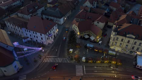 Christmas-lights-ignite-on-small-town-square,-dusk-to-night-aerial-timelapse-of-Xmas-market,-cars-pass-by-on-street,-steady-shot