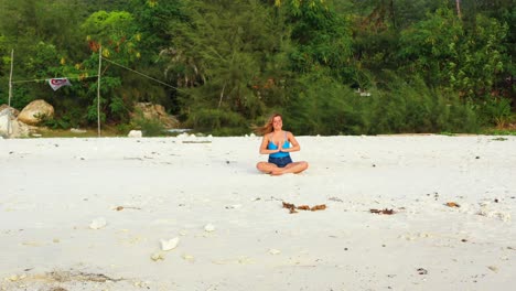 Young-woman-concentrating-on-Yoga-exercises-sitting-on-white-sand-of-exotic-beach-in-front-with-sea-on-a-windy-day