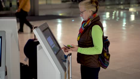 High-shot-of-woman-with-passport-and-smartphone-in-hand,-at-an-airport-self-check-in-terminal-and-checking-herself-in