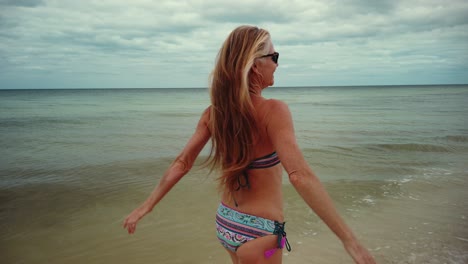 Laughing,-playful-mature-woman-in-bikini-and-sunglasses-turns-around-and-runs-on-the-beach