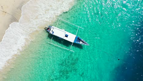 A-small-boat-sitting-on-the-surface-of-bright-blue-waters,-anchored-to-the-white-sandy-shore-of-a-tropical-beach-in-Indonesia,-Top-view