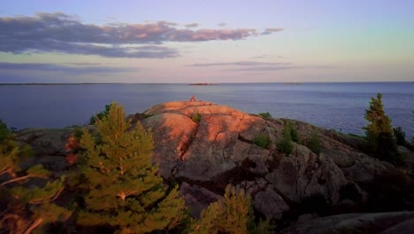 Inukshuk-on-Rocky-Pine-Tree-Island-in-Big-Blue-Lake-at-Sunset,-Drone-Aerial-Wide-Dolly-In