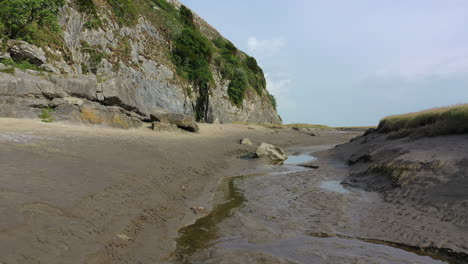 Low-moving-shot-along-the-sand-at-the-bottom-of-a-cliff,-at-low-tide-on-a-bright-day