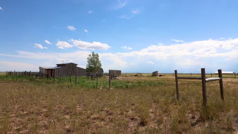 An-old-time-western-barn-with-a-corral-and-weather-vein-stands-century-over-a-barren-plain-while-puffy-white-clouds-form-and-dissipate-against-a-stark-blue-sky