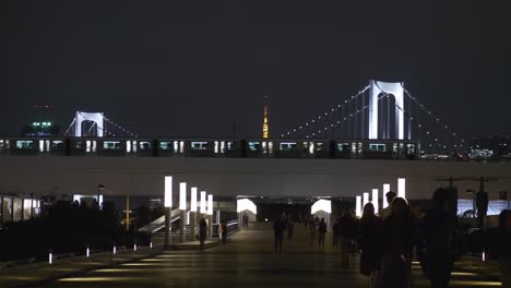 Night-scene-of-Odaiba-walkway-with-people-walking-by,-and-a-Monorail-car-passing-with-the-Rainbow-Bridge-in-the-background-in-Tokyo-Bay,-Tokyo,-Japan