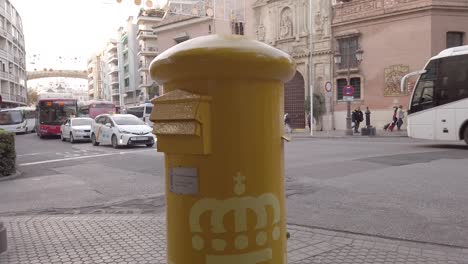 Tilt-Down-of-Yellow-Correos-Mailbox-or-Pillar-Box-in-Spain-with-Slowmo-Traffic