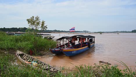 Barcos-De-Turismo-En-El-Río-Mekong-Amarrados-De-Forma-Segura-Mientras-El-Agua-Fangosa-De-La-Inundación-Se-Mueve-Río-Abajo-Mientras-Las-Banderas-Tailandesas-Ondean-Como-Se-Ve-En-La-Tarde-En-Sam-Pan-Bok-En-Ubon-Ratchathani,-Tailandia