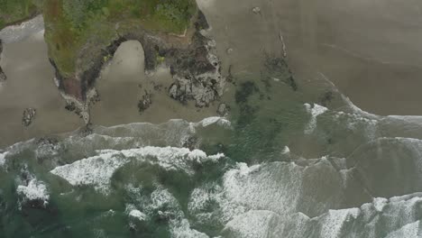 Stationary-overhead-drone-shot-of-waves-coming-into-rocky-cliffs-and-sandy-beach,-Oregon-coast