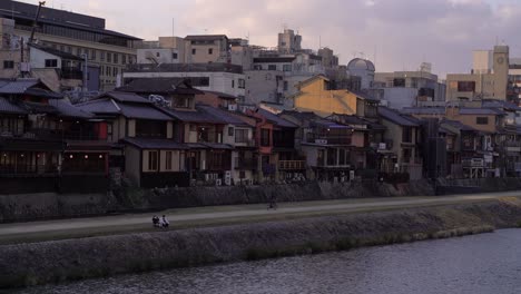 Lovely-Couple-Sitting-On-The-Riverbank-In-Kyoto,-Japan-With-Traditional-Houses-In-The-Background---Wide-Shot