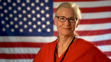 Mittelstarkes-Porträt-Einer-Krankenschwester-Im-Gesundheitswesen,-Die-Ihren-Kopf-Dreht-Und-Mit-Unscharfer-Amerikanischer-Flagge-Lächelt