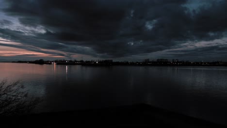 Pan-right-view-of-dark-clouds-over-still-river-waters-with-dramatic-clouds-and-colours-in-the-sunset-sky