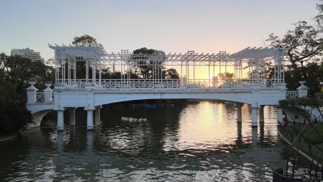 Aerial-dolly-left-of-white-bridge-over-pond-at-Rosedal-gardens-in-Palermo-neighborhood-at-golden-hour,-Buenos-Aires