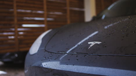 Slow-close-pan-of-Tesla-logo-on-hood-of-wet-dark-car-in-home-garage